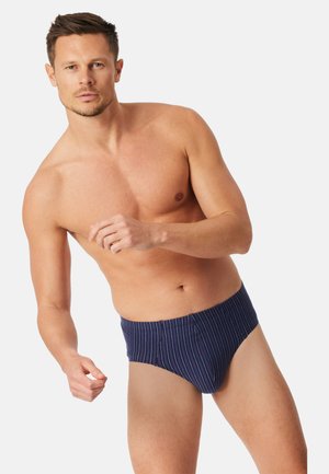 Oberkörperfreier Mann in dunkelblauen Slips mit dünnen vertikalen Streifen, posiert mit leicht nach vorne gebeugten Armen vor einem einfachen hellen Hintergrund.