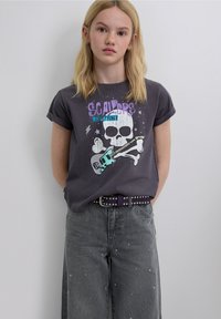 Camiseta gráfica gris de manga corta con diseño de calavera, detalles en púrpura y verde azulado, combinada con jeans grises de tiro alto con tachuelas.