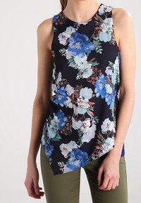 Top senza maniche a fiori con sfondo blu navy e grandi fiori blu e bianchi, orlo inclinato e tessuto leggero. Indossato con pantaloni verde oliva.