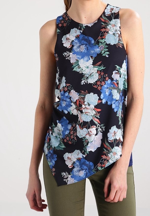 Top senza maniche a fiori con sfondo blu navy e grandi fiori blu e bianchi, orlo inclinato e tessuto leggero. Indossato con pantaloni verde oliva.