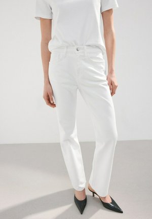 Mujer con vaqueros blancos de pierna recta, una camiseta blanca de manga corta y mules negras de tacón con punta afilada, de pie sobre un suelo claro.