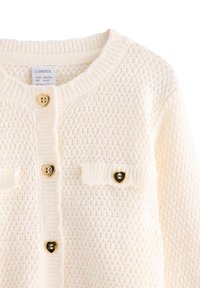 Gebreide crèmekleurige cardigan met een textuurpatroon, uitgerust met gouden hartvormige knopen en twee borstzakken met soortgelijke knoopaccenten.