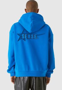 Modrá mikina s kapucňou z mäkkej látky s veľkým čiernym potlačeným textom "SENSE." a hviezdnou grafikou na chrbte. Klokaní vrecko a nastaviteľná kapucňa.