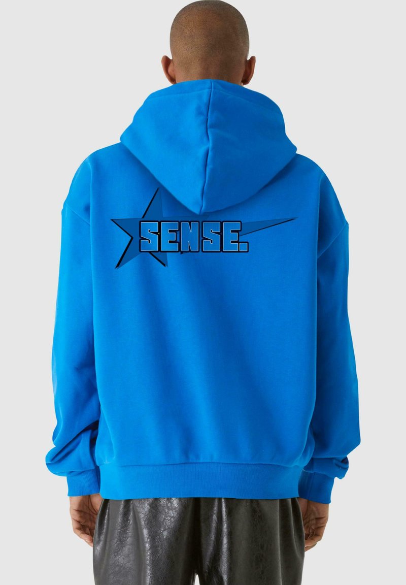 Modrá mikina s kapucňou z mäkkej látky s veľkým čiernym potlačeným textom "SENSE." a hviezdnou grafikou na chrbte. Klokaní vrecko a nastaviteľná kapucňa.