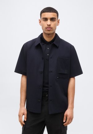Jeune homme aux cheveux courts portant une chemise noire à manches courtes boutonnée par-dessus un polo noir et un pantalon noir, sur un fond uni.