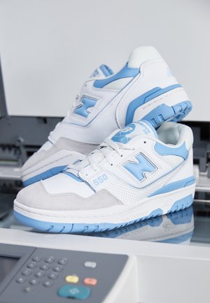 Witte en lichtblauwe New Balance 550 sneakers op het glazen oppervlak van een kopieerapparaat met in de voorgrond een zichtbaar toetsenbord.