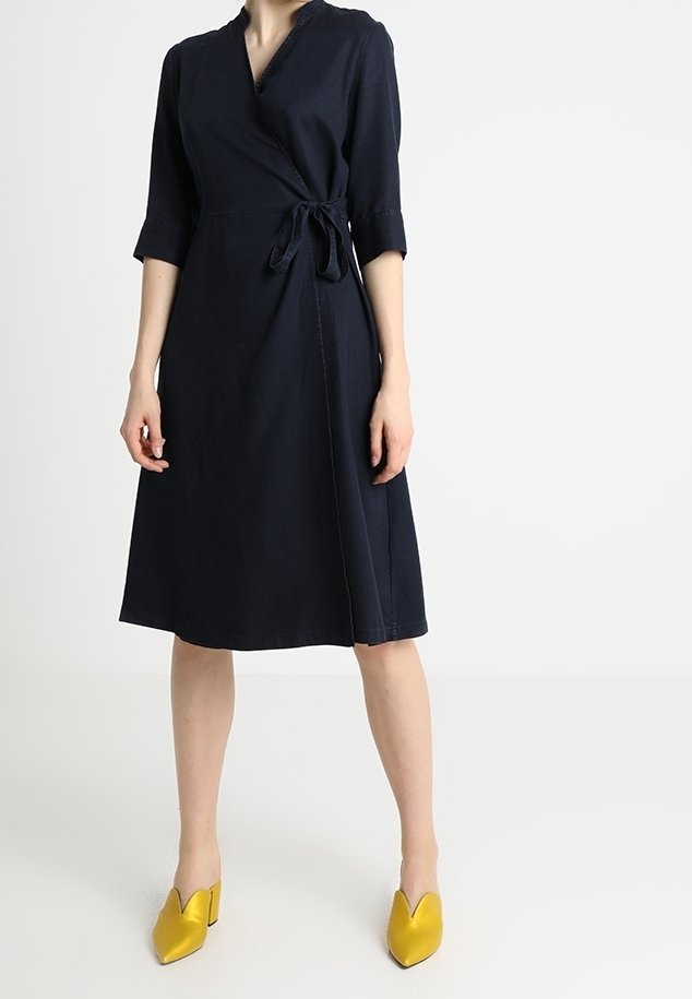 Robe cache-cœur marine avec des manches trois-quarts, détail de ceinture à nouer et longueur au niveau du genou. Associée à des chaussures à talons jaunes ornées d'un design en forme de cœur.