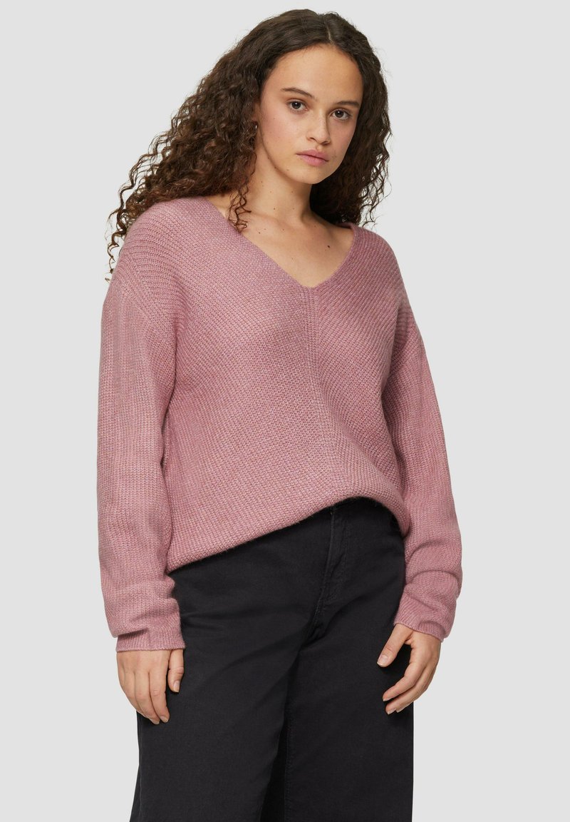 Lichtroze gebreide trui met een V-hals, lange mouwen en een ontspannen pasvorm, gecombineerd met zwarte wijduitlopende broek. Getextureerde stof, minimale details.