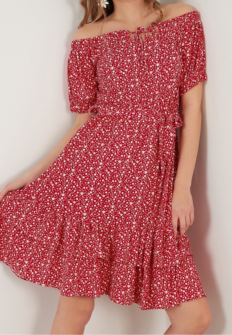 Femme portant une robe rouge à épaules dénudées avec un imprimé floral blanc, des manches bouffantes et un col noué, tenant la jupe d'une main.