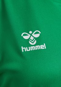 Zielona sportowa tkanina o teksturowanym wykończeniu z białym logo Hummel, zaprojektowanym z biało-żółtym emblematem przypominającym pszczołę i stylizowanymi elementami.