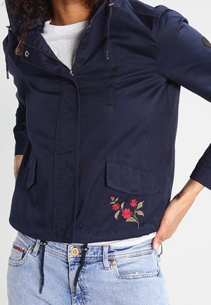 Granatowa kurtka z przednimi kieszeniami i czerwonym kwiatowym haftem na jasnoszarej koszuli oraz jasnoniebieskich dżinsach.