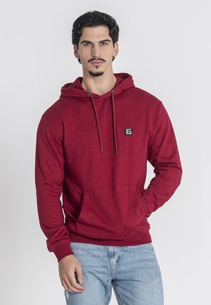 Rød hættetrøje lavet af blødt stof, med en frontlomme, justerbar snører og et lille logo-patch på brystet.