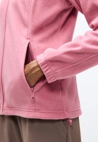 Χέρι σε τσέπη με φερμουάρ ροζ ζακέτας από φλις με ελαστικό τελείωμα, συνδυασμένη με καφέ παντελόνι πάνω σε απλό φόντο.