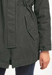 Giacca leggera verde scuro con zip frontale, vita elastica, tasca laterale e polsini con bottoni. Il tessuto testurizzato completa il look.
