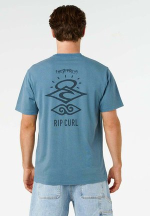 Man draagt een blauw Rip Curl T-shirt met een zwarte afbeelding en de tekst "The Search" op de achterkant, gecombineerd met lichtblauwe denim shorts.