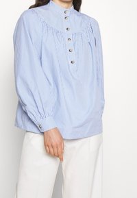 Blusa a righe blu chiaro e bianco con collo alto, dettagli di bottoni e maniche arricciate. Il tessuto appare morbido e leggero.