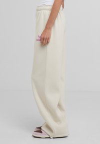 Pantaloni della tuta beige con vita elasticizzata, gambe larghe e testo ricamato rosa. Il tessuto ha una consistenza morbida e liscia.