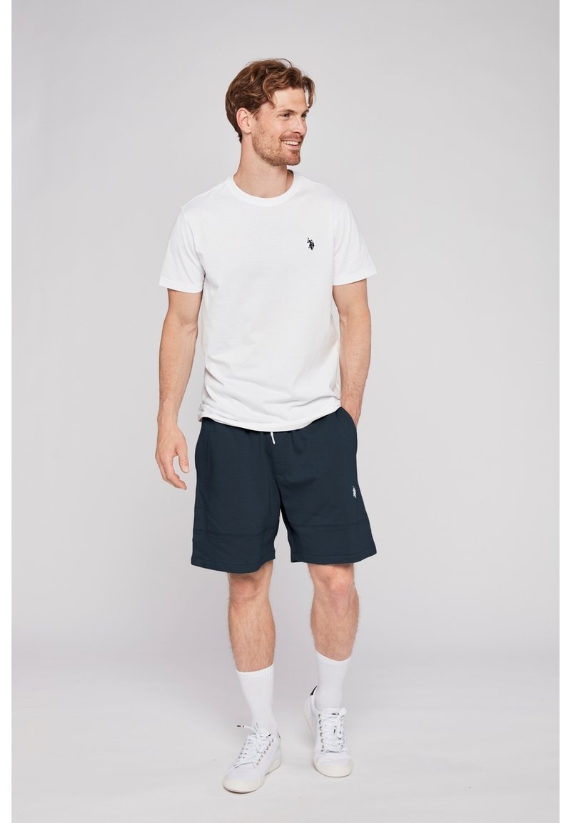White short-sleeve t-shirt with a small black logo, paired with navy blue shorts and white sneakers, accentuated by white socks.