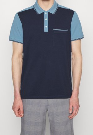 Man wearing a navy blue and light blue color-block polo shirt with short sleeves and a chest pocket, paired with gray plaid pants.