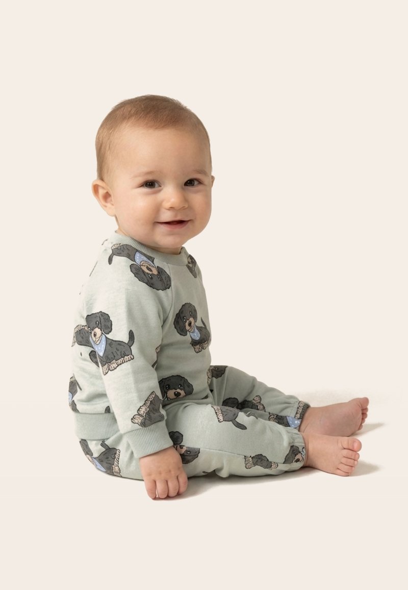 Bébé souriant assis pieds nus, portant un pyjama vert clair avec des illustrations de chiens noirs et des bandanas bleus sur un fond neutre.