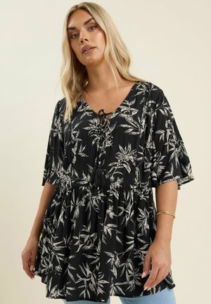 Blonde vrouw die een losse zwarte tuniek draagt met witte bloemenprint, korte brede mouwen en een strik met franjes bij de halslijn.