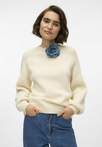 Maglione lavorato a maglia color avorio con maniche a coste e scollo tondeggiante, decorato con una spilla a forma di fiore in denim sul lato sinistro. Abbinato a jeans blu.