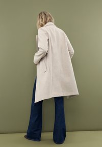 Beige uldblandingsfrakke med en notch-krave og sidelommer, parret med mørkeblå flagrende jeans. Baggrunden er en dæmpet olivengrøn.