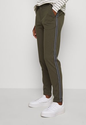 Personne portant un pantalon vert olive avec des bandes latérales noires et blanches et des baskets blanches, debout les mains dans les poches.