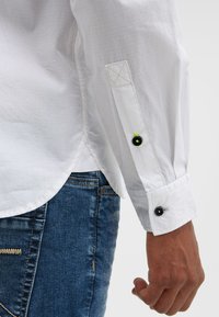 White button-up shirt with adjustable cuffs, featuring a black button and green accent. Worn with blue denim jeans, showing textured fabric.