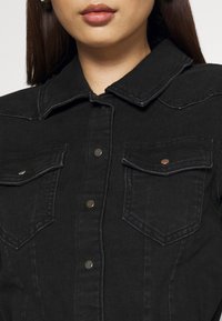 Primer plano de una persona que lleva una chaqueta de mezclilla negra con botones a presión y dos bolsillos en el pecho con solapas.