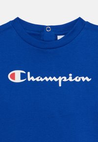 T-shirt in cotone blu con colletto rotondo, con logo "Champion" bianco e un piccolo dettaglio a forma di anello sulla parte posteriore del collo.