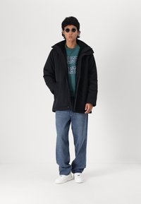 Black padded jacket with a high collar, worn over a green sweatshirt featuring white text. Complemented by light blue jeans and white sneakers.