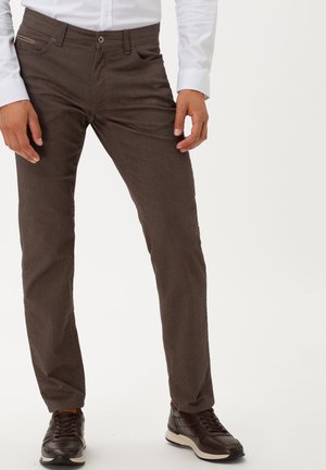 Homme portant un pantalon texturé marron foncé, des baskets marron foncé à lacets, et une chemise blanche boutonnée, debout devant un fond uni.