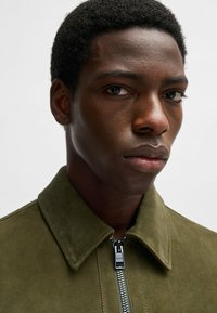 Olivgrön mockajacka med dragkedja fram, med spetsig krage och synlig sömnad. Slät textur och metalldragkedjedetalj.