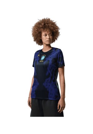 Femeie cu păr creț, purtând un tricou de fotbal al Braziliei, negru și albastru, cu stema echipei și cinci stele, stând pe un fundal alb.