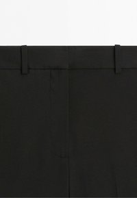 Close-up van de tailleband van een zwarte pantalon met riemlussen en detail van de voorste naad.