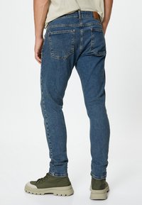Vaqueros de mezclilla azul ajustados con una textura suave, diseño de cinco bolsillos y una etiqueta de cuero marrón en la cintura. Combinados con zapatos verdes.