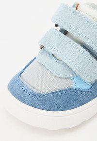 Zapatilla azul y gris con materiales de ante y malla, correas de velcro, punta redondeada y suela de goma blanca. Detalles de costura notables visibles.