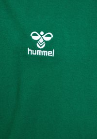 Wit geborduurd Hummel-logo, een gestileerde bij boven de merknaam op een groene stoffen achtergrond.