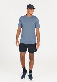 Harmaa kosteutta siirtävä urheilupaita, mustat shortsit, musta lippis ja siniset lenkkarit. Malli seisoo yksinkertaista valkoista taustaa vasten.