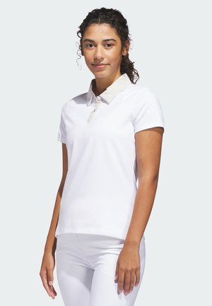 Femme aux cheveux bouclés portant un polo blanc à manches courtes et un pantalon blanc, debout devant un fond clair.