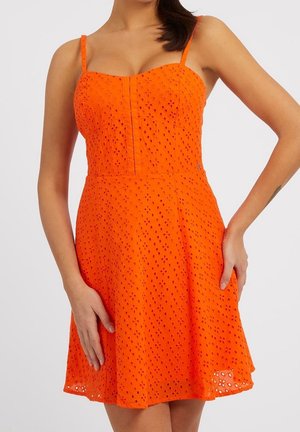 Femme portant une robe orange sans manches avec un motif œillet et un corsage ajusté, debout avec une main sur la hanche et l'autre posée vers le bas.