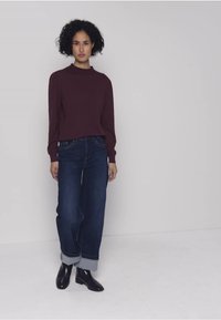 Bordeauxroter Pullover mit langen Ärmeln und hohem Kragen. Blaue Jeans mit geradem Bein und umgeschlagenen Säumen. Schwarze Stiefeletten. Schlichtes, sauberes Design ohne Muster.