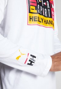 Cămașă albă cu mânecă lungă, având un imprimeu grafic îndrăzneț cu un amestec de roșu, galben și negru. Include branding pe mânecă cu un logo.