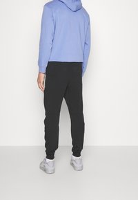 En kortärmad ljusblå hoodie med huva, tillsammans med svarta smala joggers. Grå sportskor synliga, stående på en enfärgad bakgrund.