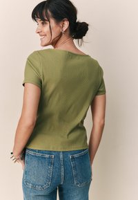 Oliivivihreä ribbed lyhyet hihatoppi, jossa on leveä kaula-aukko, yhdistettynä korkeavyötäröisiin sinisiin farkkuihin, joissa on kaksi takataskua.