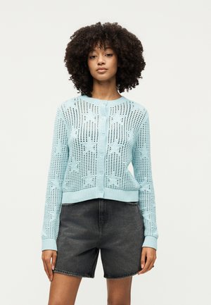 Göndör hajú nő világoskék kötött kardigánt visel csillagmintákkal és fekete farmer rövidnadrágot, egyszerű világos háttér előtt áll.