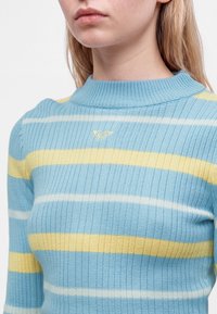 Femme portant un pull côtelé bleu avec des rayures horizontales jaunes et blanches et un petit logo brodé sur la poitrine.