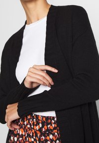 Svart stickad kofta med ribbade detaljer, öppen framsida och långa ärmar, bärs över en vit topp och ett mönstrat kjol i orange och svart.