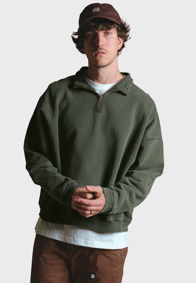 Sudadera de forro polar verde oliva con cuello de media cremallera, presenta un corte relajado, puños acanalados y una camiseta blanca en capas debajo.